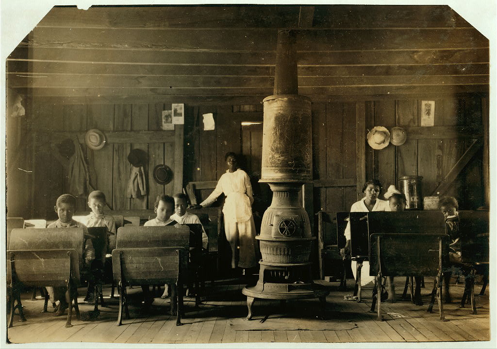 A black-and-white image of a small room with one African-American teacher and six students in a small room with a woodburning stove in the center of the room.